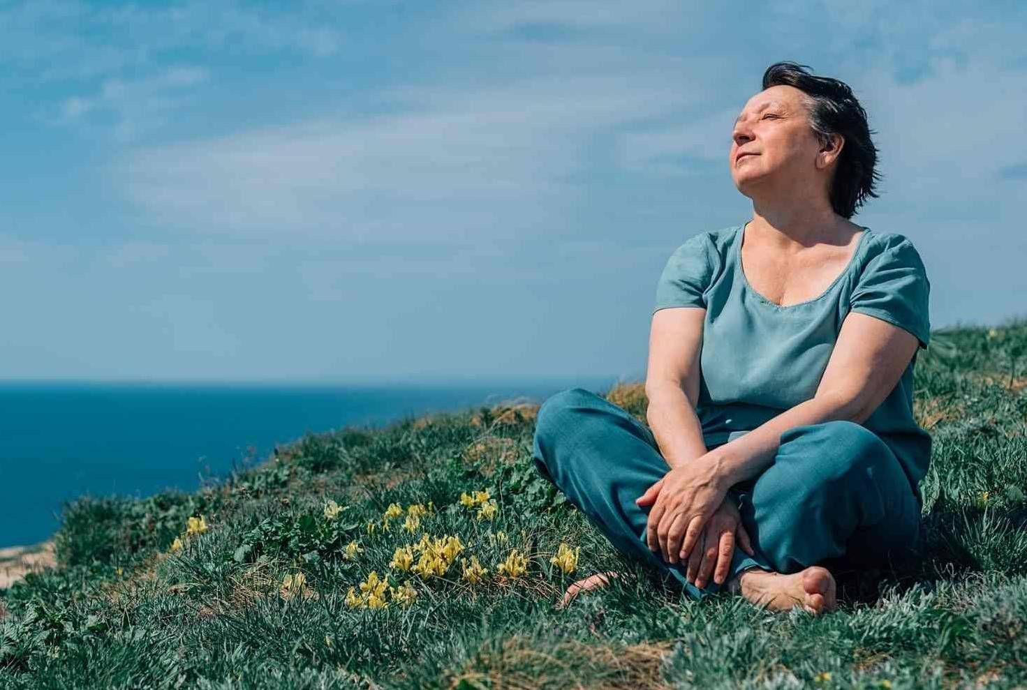Una persona sentada en un acantilado cubierto de hierba sobre el océano, mirando hacia arriba bajo un cielo azul.