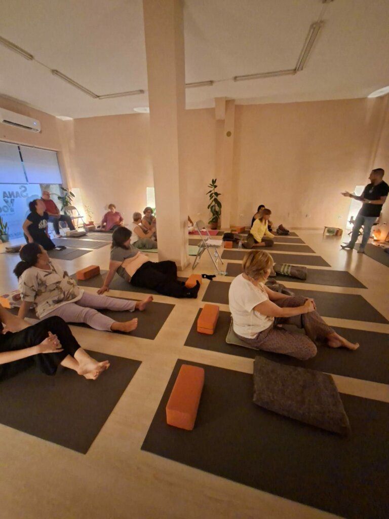 Personas practicando yoga sobre esterillas en un estudio con iluminación tenue, mientras un orador dirige la sesión.