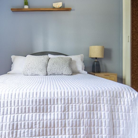Neatly made bed with white quilt in a light gray bedroom, with pillows, bedside lamp, and wall shelf.