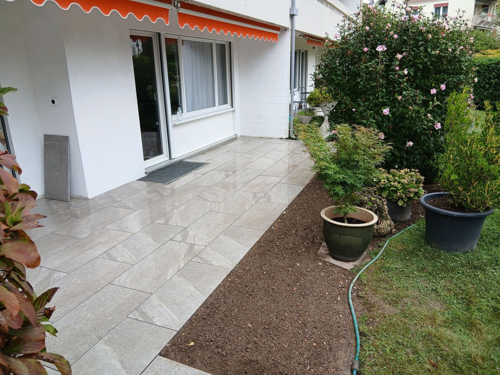 Eine Steinterrasse neben einem weissen Gebäude mit orangefarbener Markise, umgeben von einem Gartenbeet mit Topfpflanzen und Rasen.