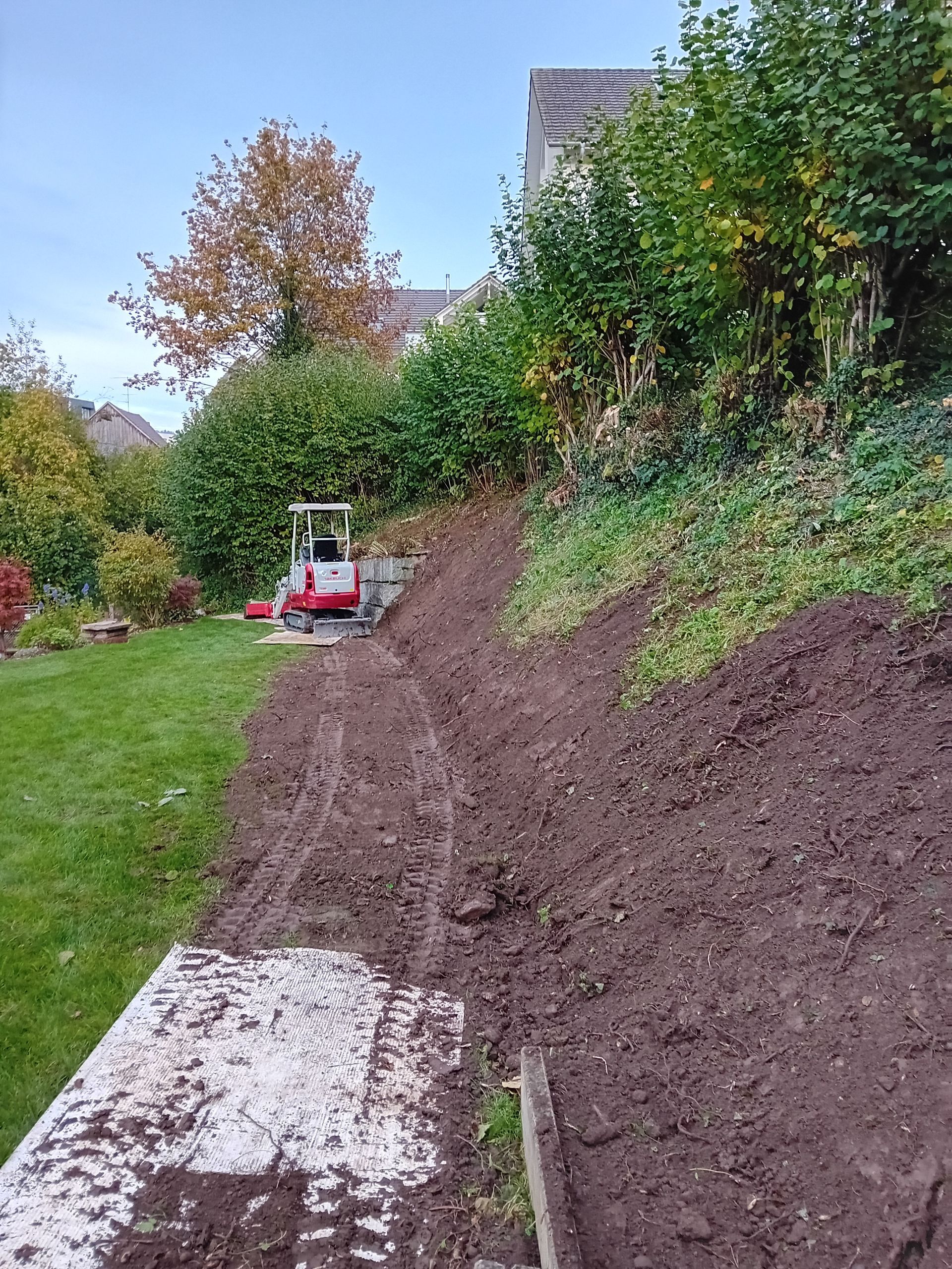 Ein kleiner Bagger steht in einem schlammigen, teilweise ausgehobenen Gartenbeet unterhalb eines mit Bäumen und Sträuchern bewachsenen Hangs.
