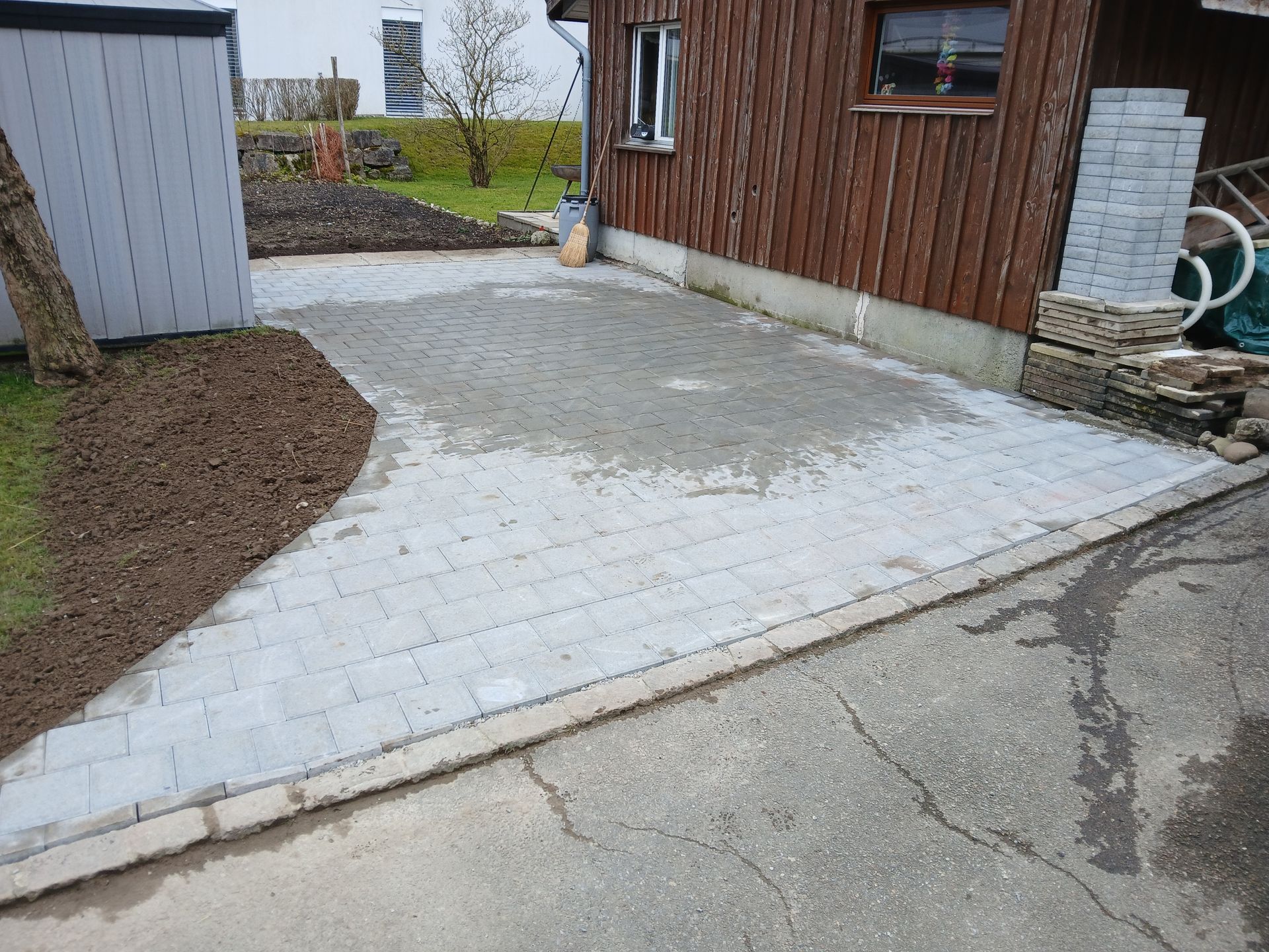 Eine teilweise fertiggestellte, hellgraue Steinpflasterauffahrt neben einem Holzgebäude und einem Gartenbeet.