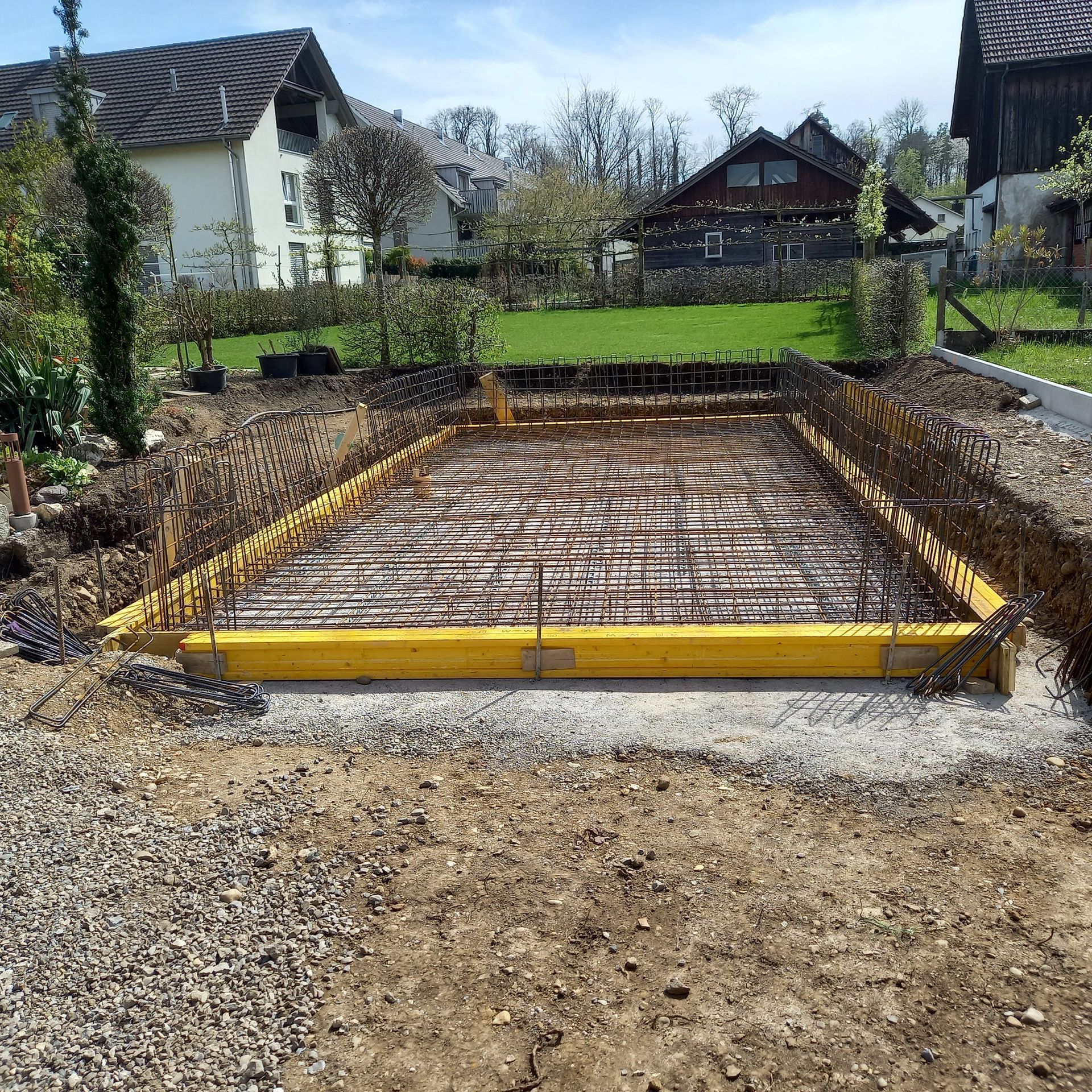 Eine rechteckige, mit gelben Holzschalungen und Stahlbewehrungsmatten ausgekleidete Betonfundamentgrube in einem Wohngarten.