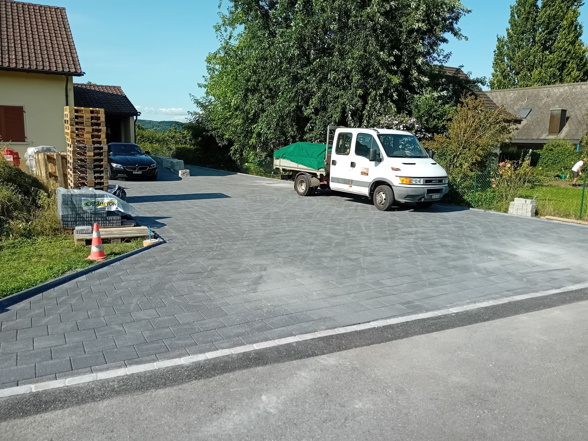 Ein weisser Lastwagen mit einer grünen Plane parkt auf einem gepflasterten Parkplatz neben einem Gebäude und einem schwarzen Auto.