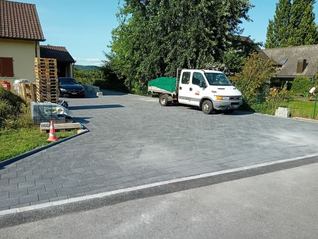 Ein weisser Lastwagen mit einer grünen Plane parkt auf einem gepflasterten Parkplatz neben einem Gebäude und einem schwarzen Auto.