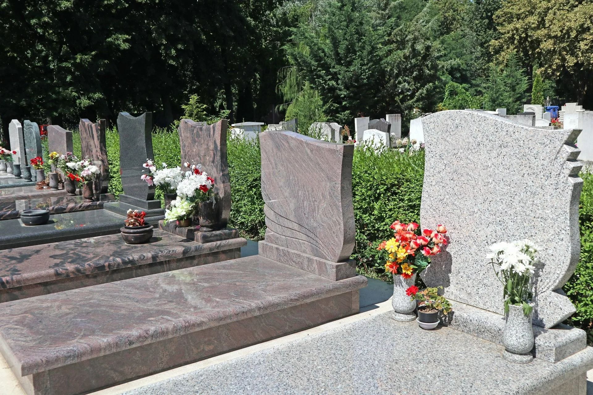 Cimetière avec plusieurs pierres tombales, certaines fleuries, arbres en arrière-plan, journée ensoleillée.