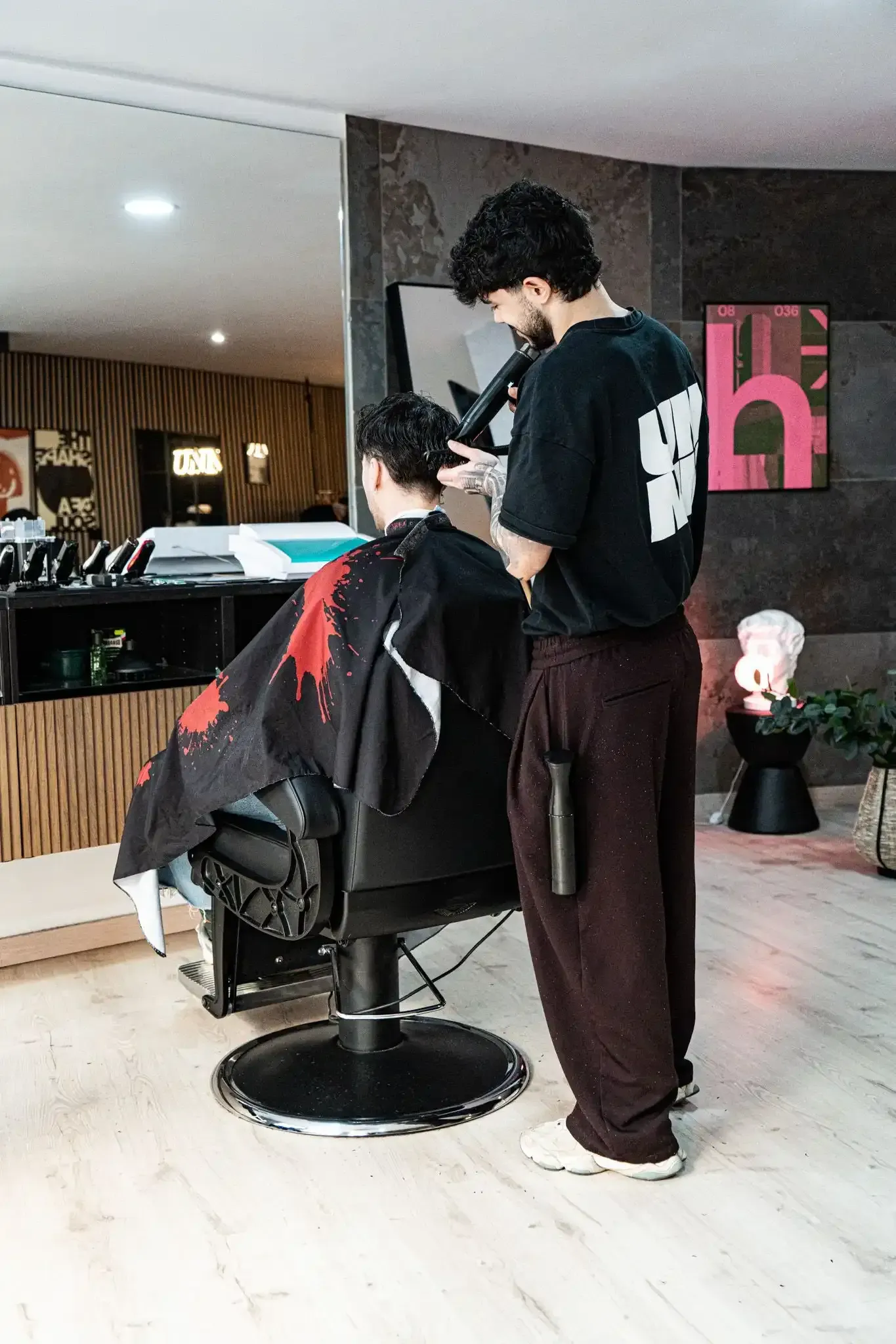 Interior de peluquería con dos sillones, una mesa pequeña y una persona entrando por la puerta. Pared con acento de madera.