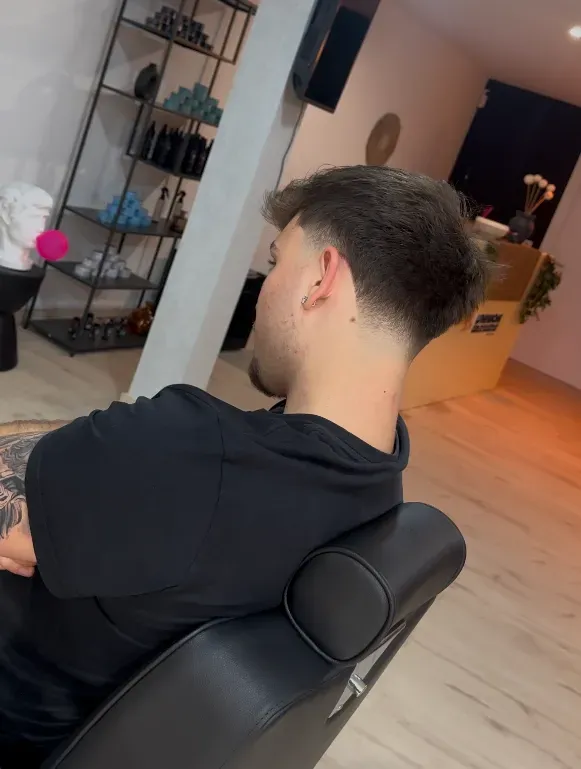 Un hombre con el pelo descolorido se sienta en una silla de barbero. Cabello oscuro, camisa negra y un ambiente de salón.