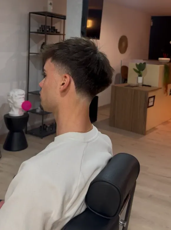 Hombre con corte de pelo degradado, sentado en una silla de peluquería. Mira a la izquierda y viste una camisa blanca.