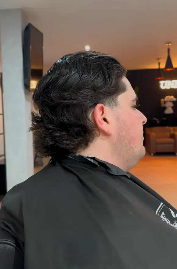 Hombre con corte de pelo; cabello oscuro con la parte superior peinada y los lados más cortos; dentro de un salón.