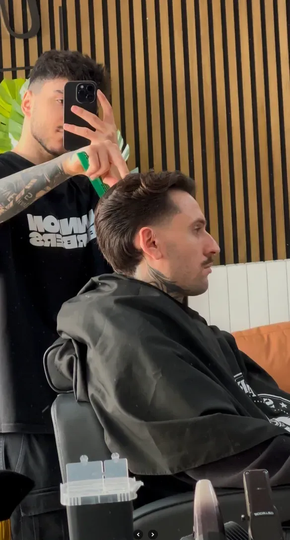 Hombre cortándose el pelo en una peluquería. Un barbero con camisa negra se toma una selfie mientras corta el pelo.