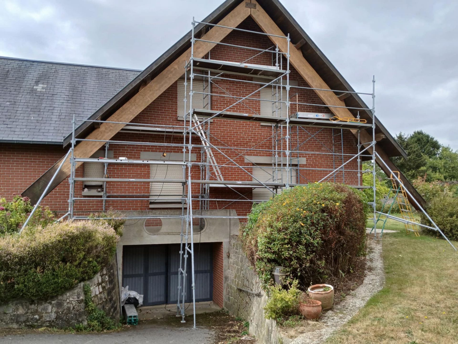 Échafaudages installés sur une maison en briques en cours de rénovation ; une grosse poutre en bois soutient le toit.