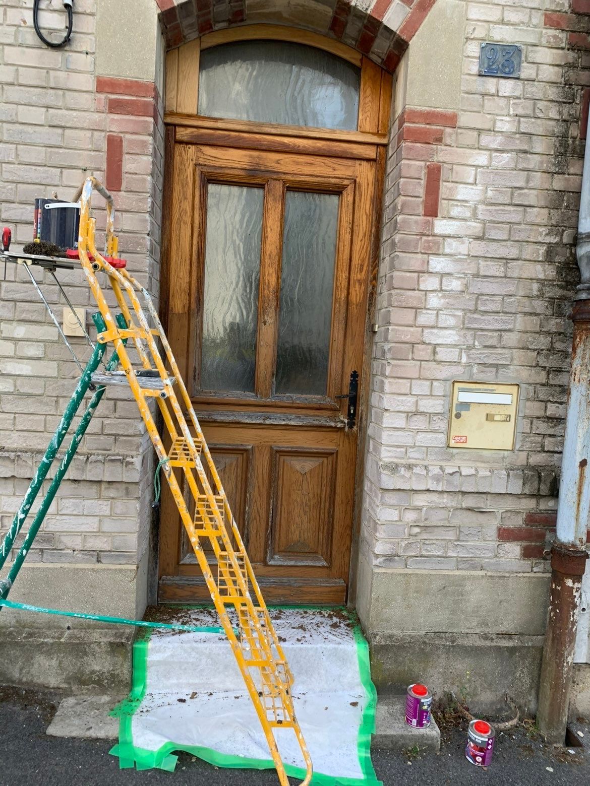Une porte en bois patinée par le temps, avec des panneaux de verre, une échelle et du matériel de peinture, sur la façade d'un bâtiment en briques.