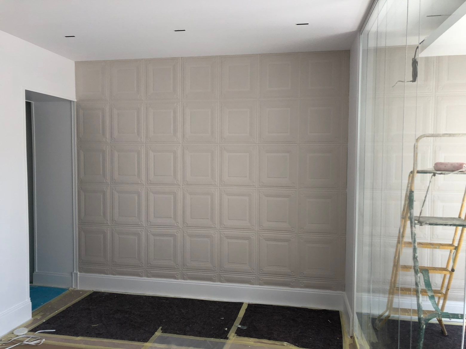Pièce avec papier peint beige à panneaux, échelle et cloison vitrée.
