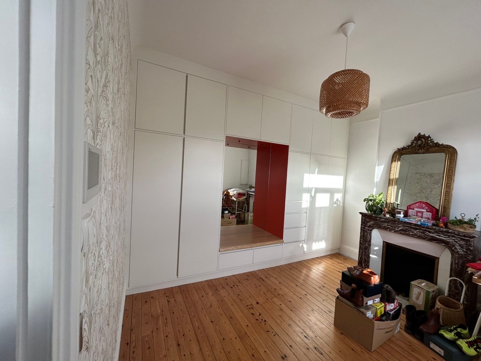 Pièce lumineuse avec rangements intégrés blancs, porte rouge, parquet, cheminée et suspension lumineuse en tissu tressé.