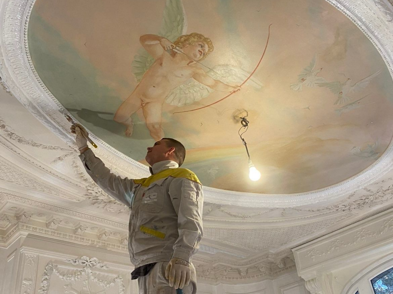 Un peintre, juché sur un échafaudage, travaille sur une fresque au plafond.