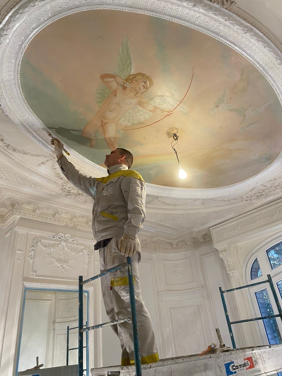 Un peintre sur un échafaudage effectue des retouches sur une fresque murale.