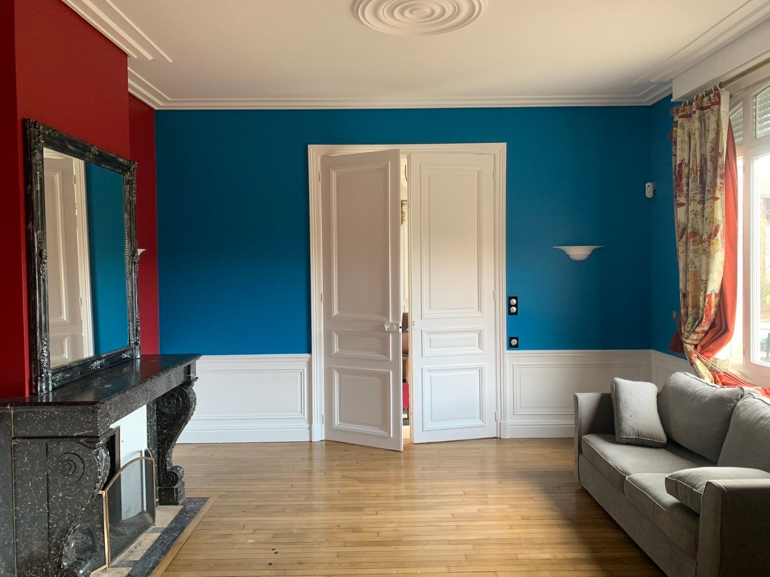Salon avec murs bleu et rouge, moulures blanches, parquet, canapé gris et cheminée ornée.
