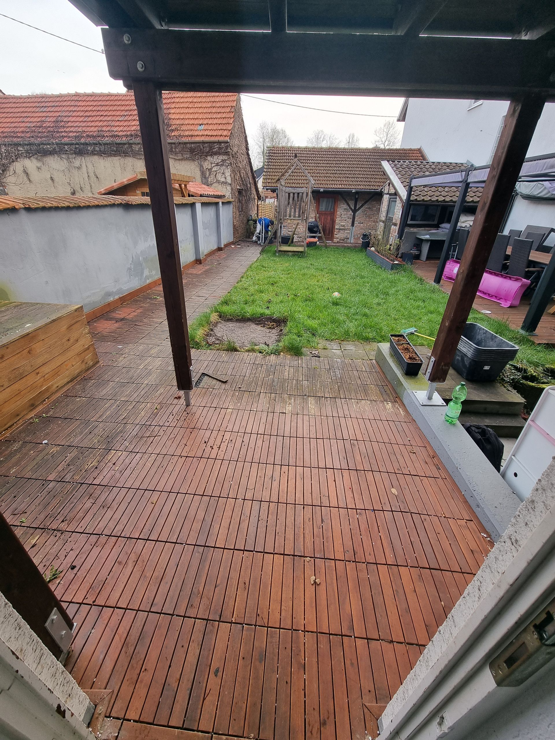 Überdachte Terrasse im Hinterhof mit braunen Terrassenfliesen, ein Rasengarten und im Hintergrund ein kleiner Schuppen.