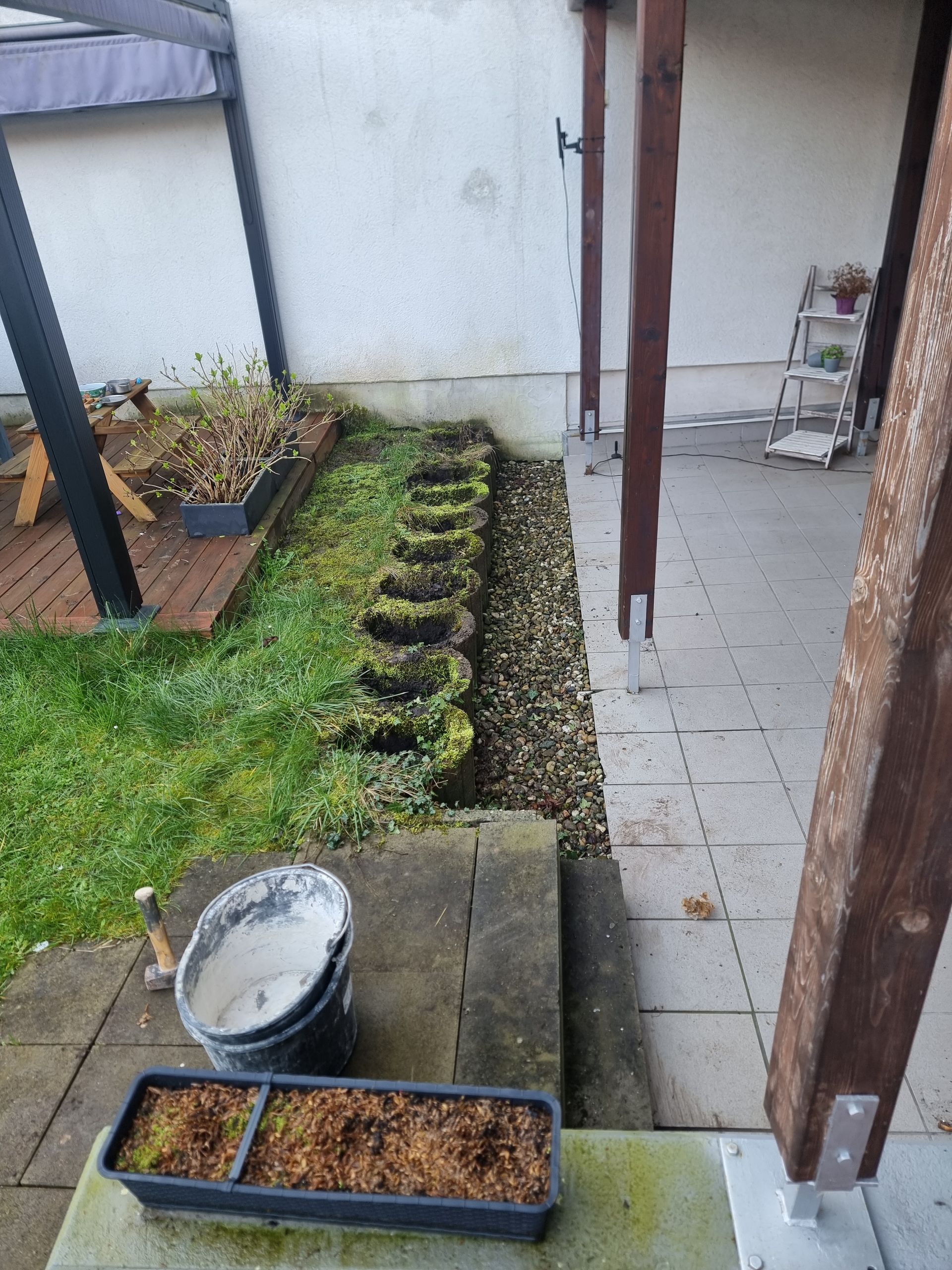 Schmale Gartenterrasse mit Topfpflanzen, gefliestem Gehweg und überdachter Veranda mit Pfosten