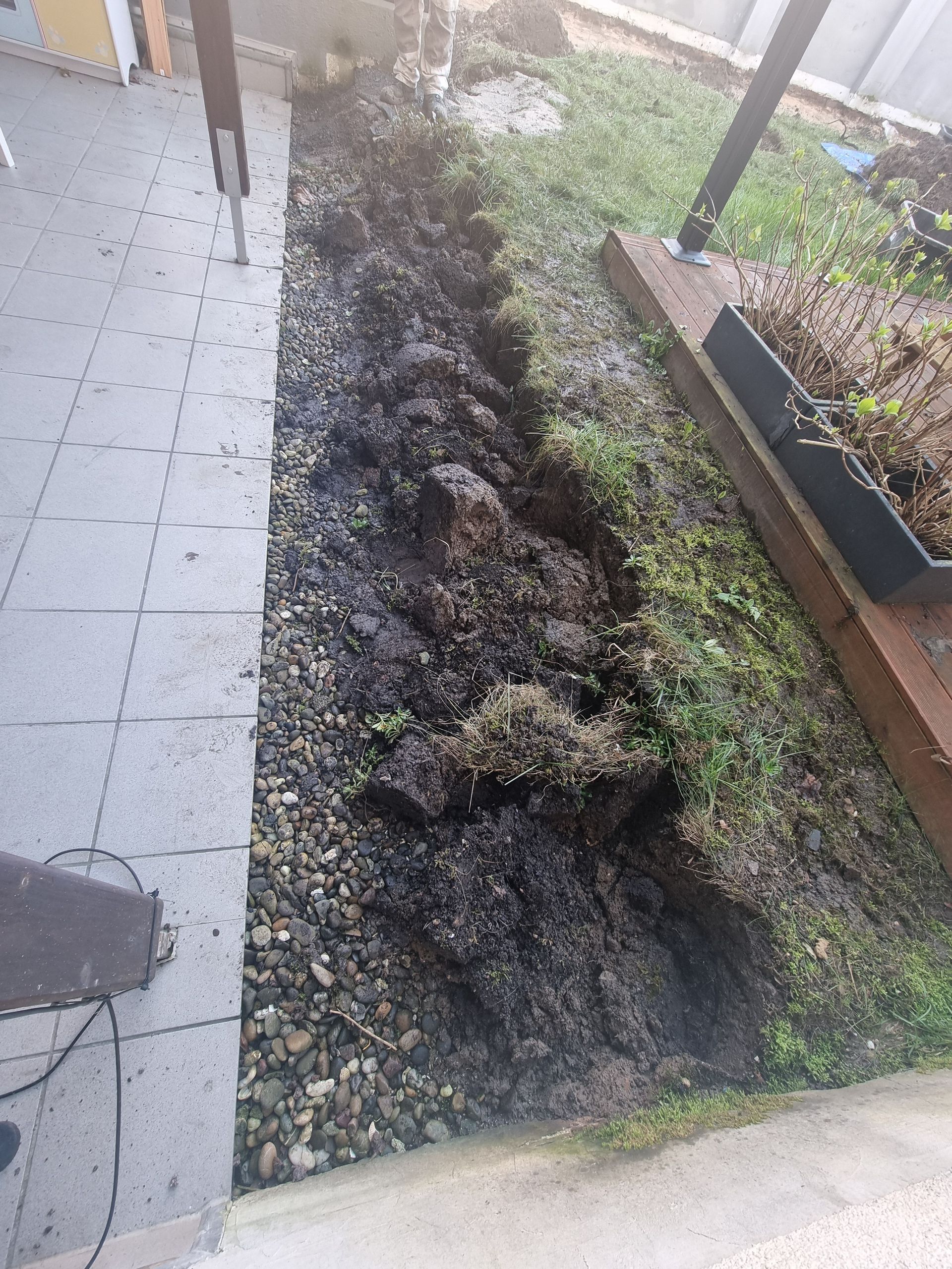 Ein frisch ausgehobener Graben neben einem gefliesten Gehweg und einem Gartenbeet, dessen Rand mit freiliegender Erde und Kies bedeckt ist.