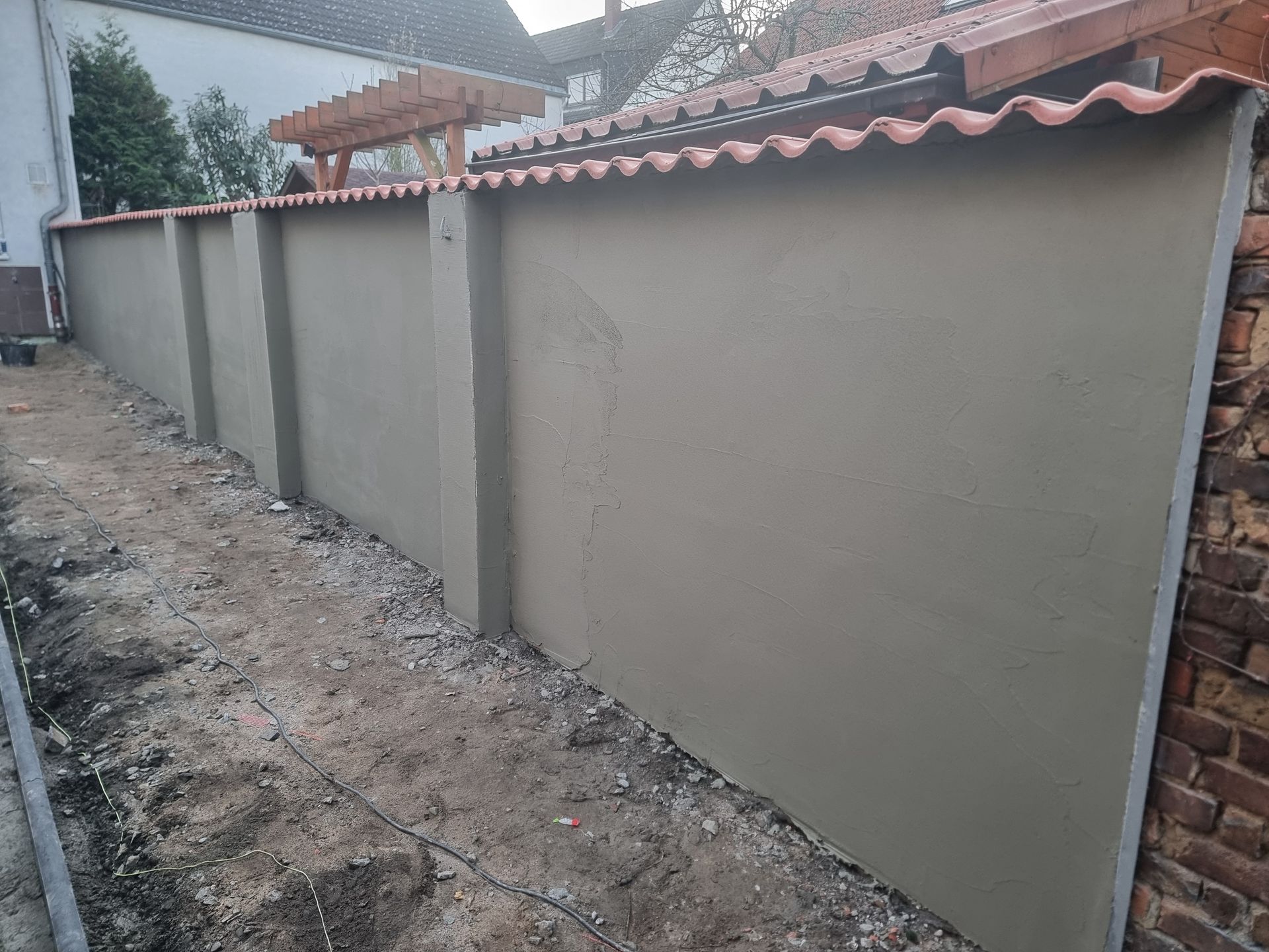 Eine frisch verputzte graue Mauer entlang eines schmalen Feldwegs neben einem Backsteinhaus unter einem roten Ziegeldach.