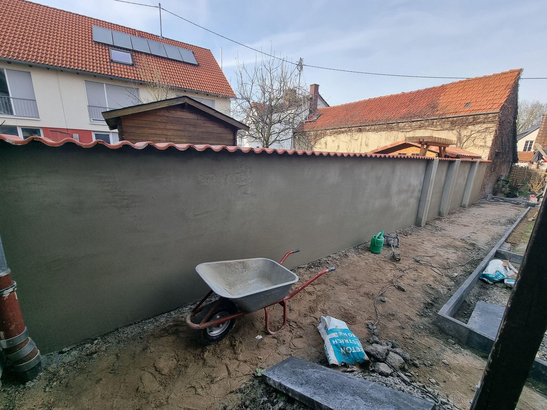 Schmaler Hinterhof mit einer Schubkarre neben einer langen grauen Mauer und Häusern mit Ziegeldächern.
