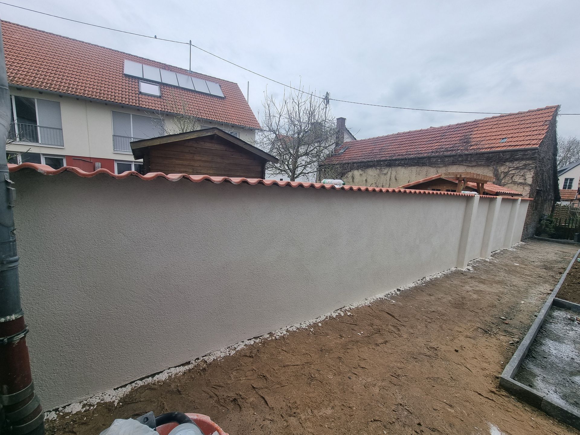 Eine lange beige Mauer neben einem Feldweg, rote Ziegeldächer und ein bewölkter Himmel in einem Wohnhof