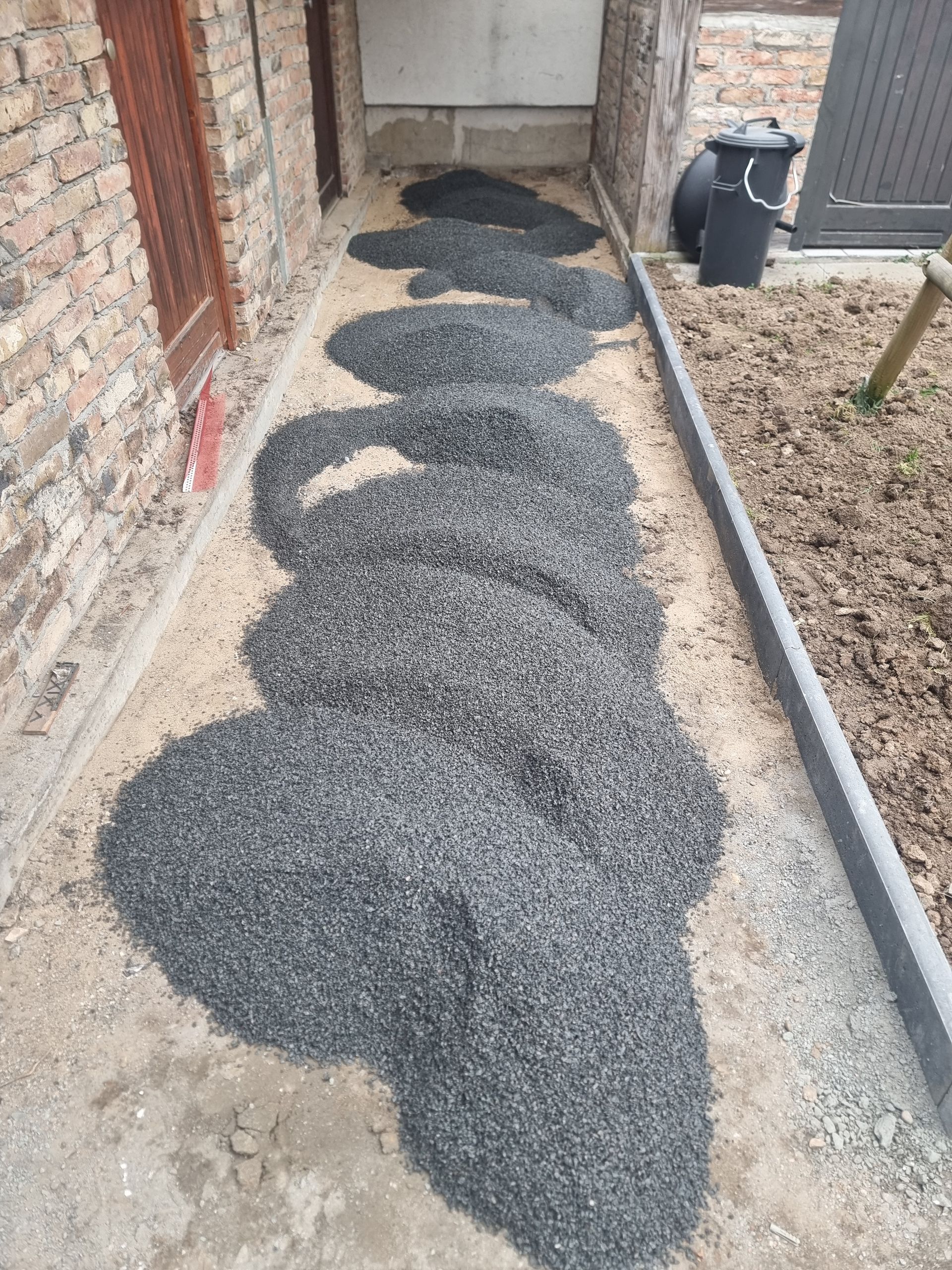 Graue Kieshaufen säumen einen schmalen Betonweg neben einer Backsteinmauer und einem Zaun.