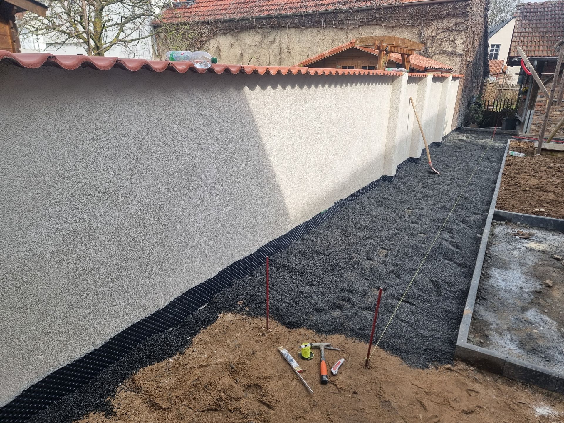Schmaler Seitenhof mit frischem Kies neben einer weißen Stuckwand und Dachlinie, der laufende Landschaftsbauarbeiten erkennen lässt.