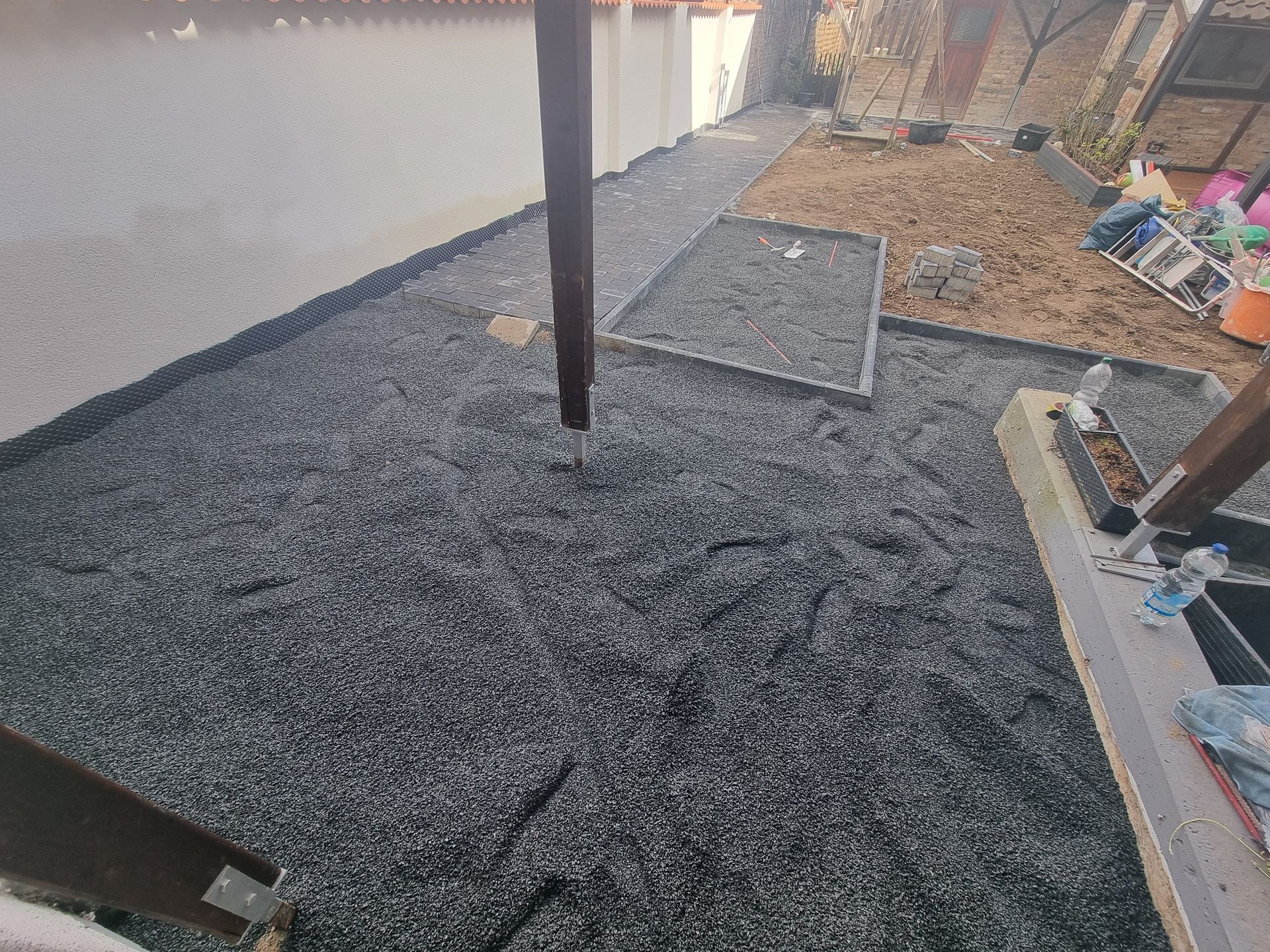 Schwarzer Kiesplatz im Bau neben einer Mauer, mit einem Mittelpfosten und Werkzeugen/Materialien an der rechten Seite.