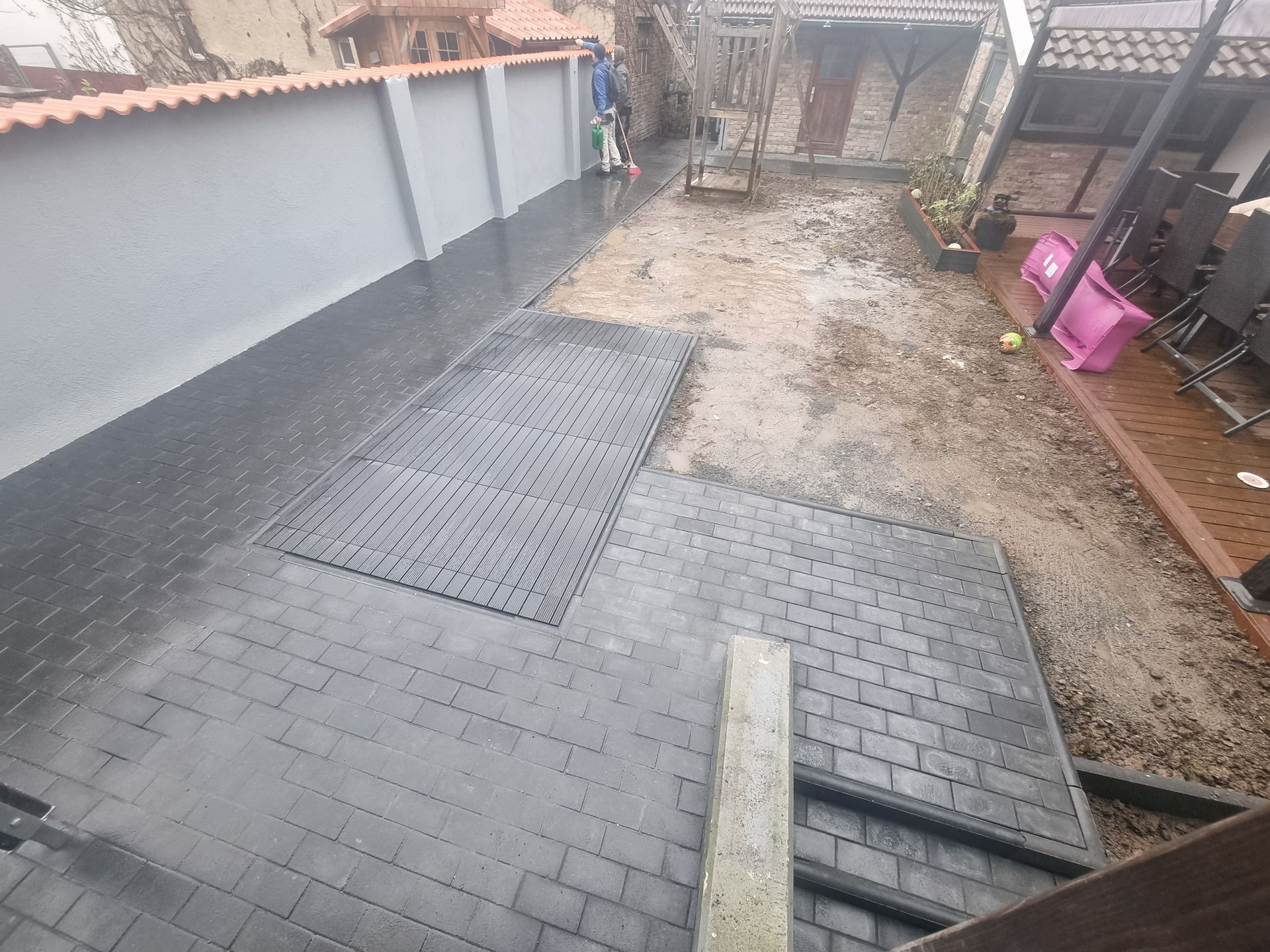 Hinterhof mit dunklem Backsteinpflaster, einem Kiesweg und einem eingezäunten Bereich mit rosa Eimern auf der rechten Seite