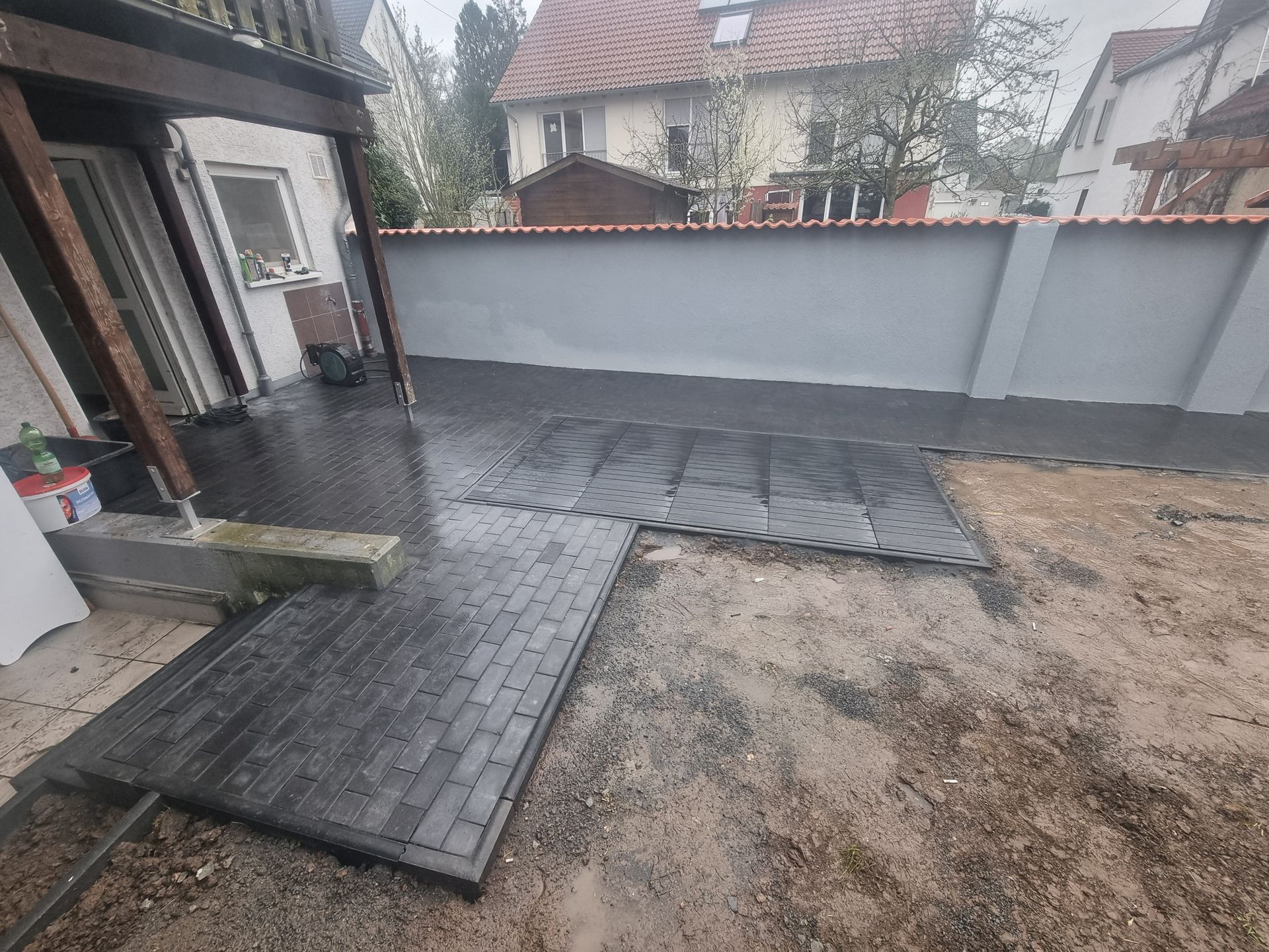 Hinterhofterrasse mit nassen Pflastersteinen, einer weißen Mauer und einem unbefestigten Fleckchen Erde neben einem Haus.