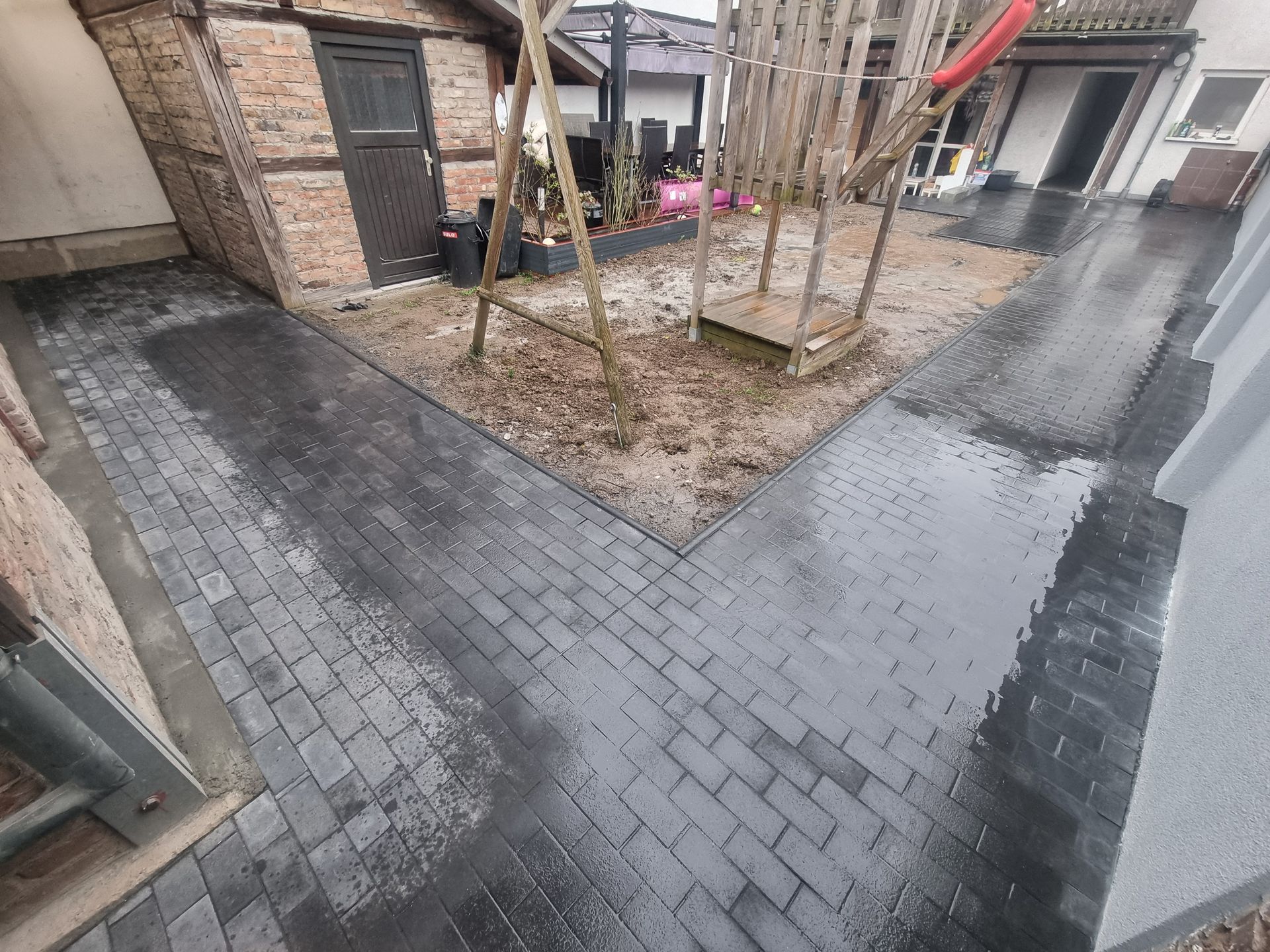 Regnerischer Innenhof mit nassen, grauen Pflastersteinen, einem kleinen Baumbeet und Hauseingängen entlang eines schmalen Weges.