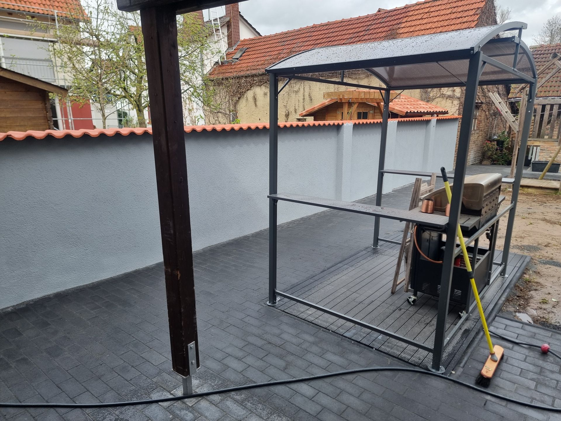 Ein Metallkäfig steht auf einer Einfahrt, daneben befinden sich ein Arbeiter und Ausrüstung neben einer grauen Mauer unter einem Carport.