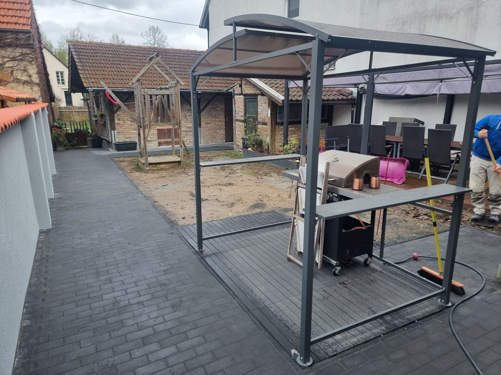 Überdachte Terrasse mit Metallrahmen, einem Tisch und Gartengeräten neben einem gepflasterten Gehweg.