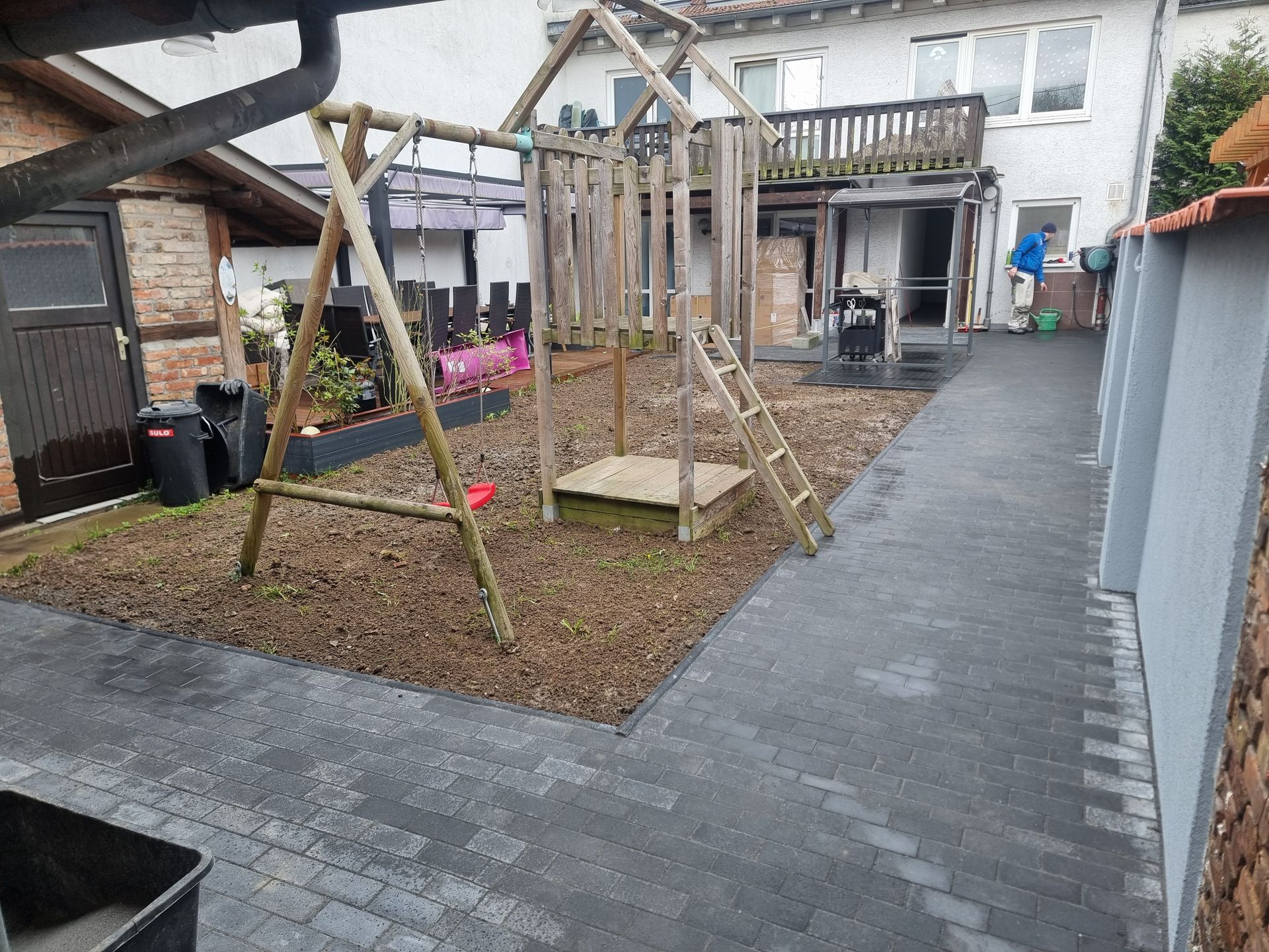 Hinterhof mit hölzernen Spielschaukeln, Kiesfläche und einem gepflasterten Weg neben einem Haus und einem Schuppen