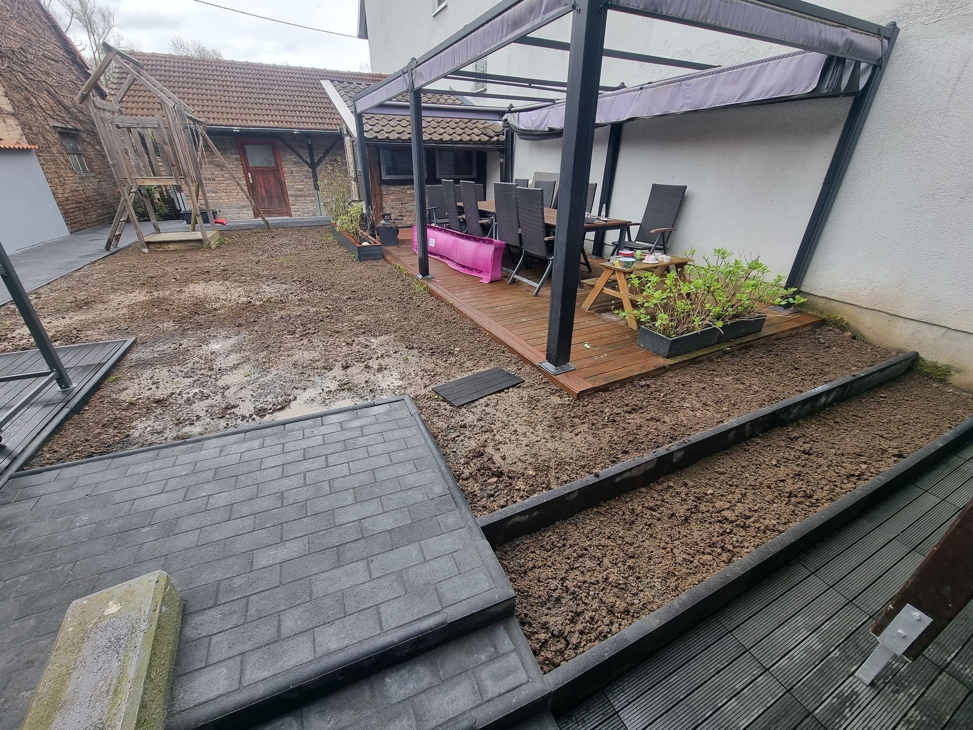Hinterhofterrasse mit grauen Pflastersteinen, Holzpergola, rosa Bank und Hochbeeten mit Mulch.