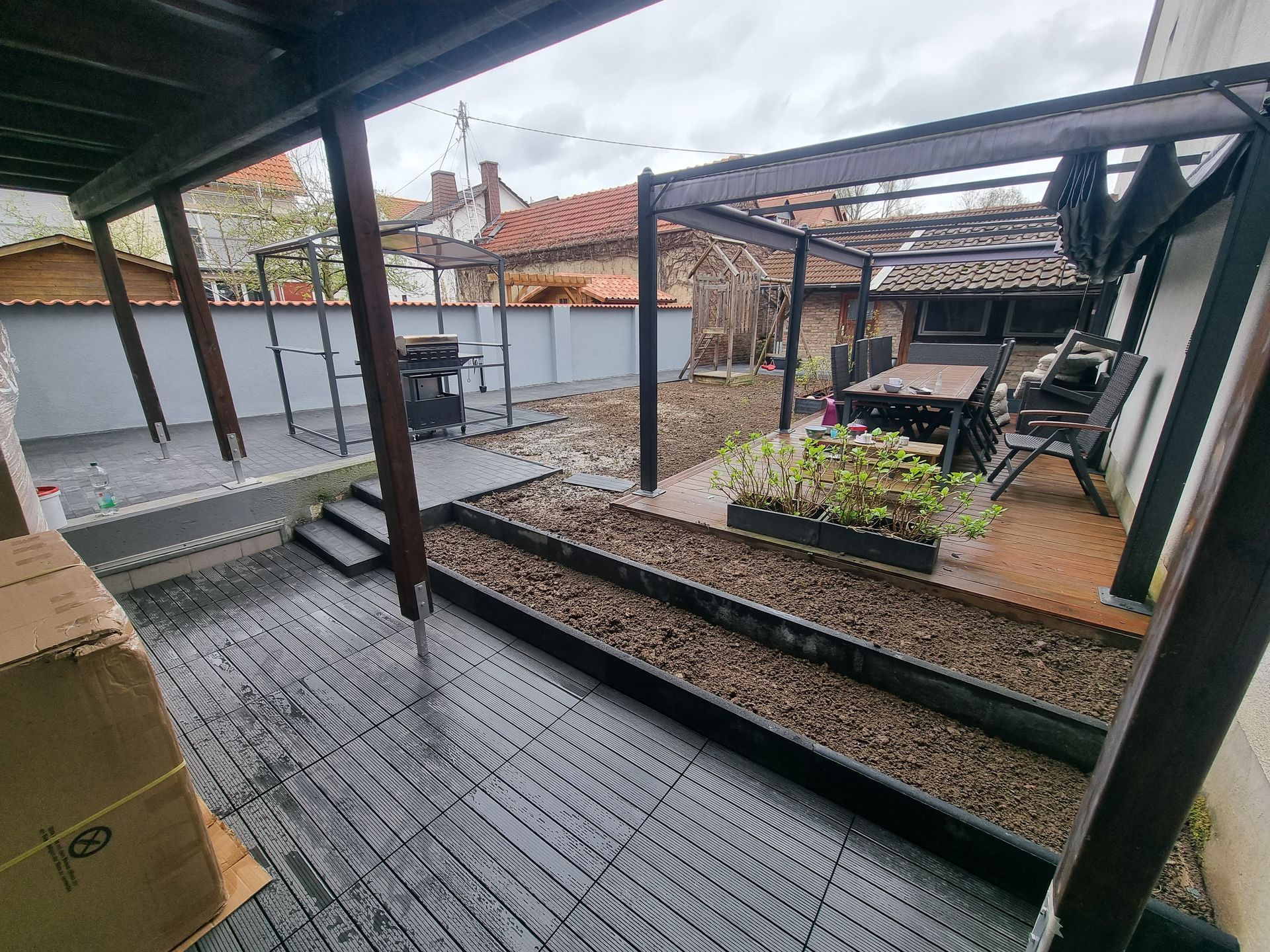 Überdachte Terrasse im Hinterhof mit Nasssteinboden, Metallpfosten und Sitzgelegenheiten im Freien neben einem kleinen Blumenbeet.