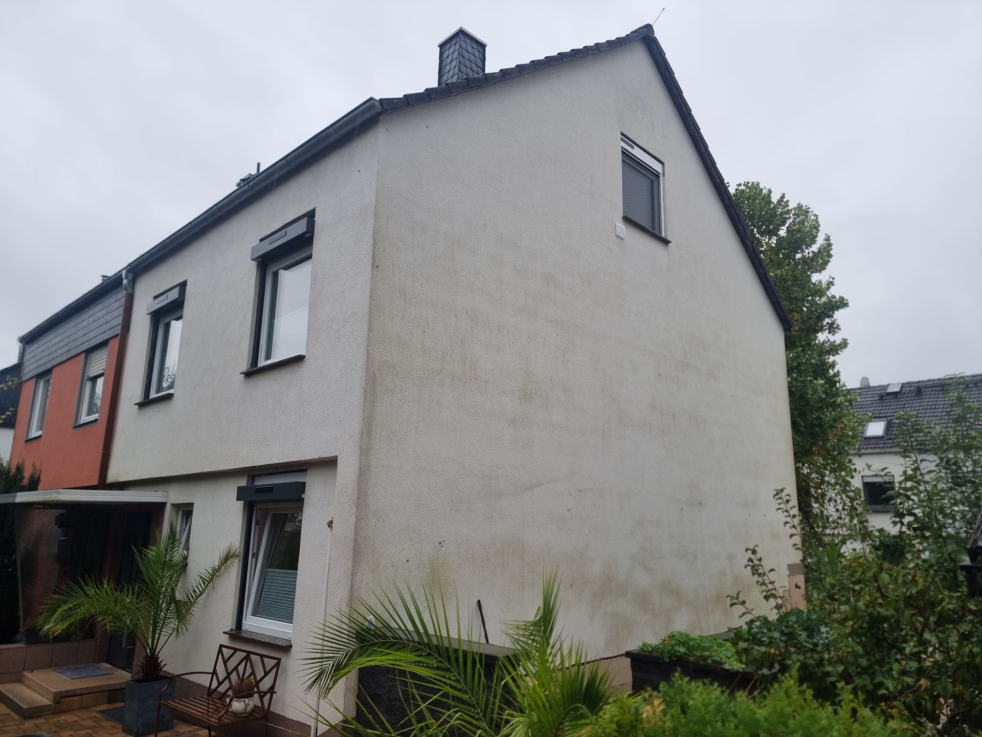 Weißes zweistöckiges Haus mit schwarz umrandeten Fenstern und einem kleinen Vorgarten neben einem roten Gebäude