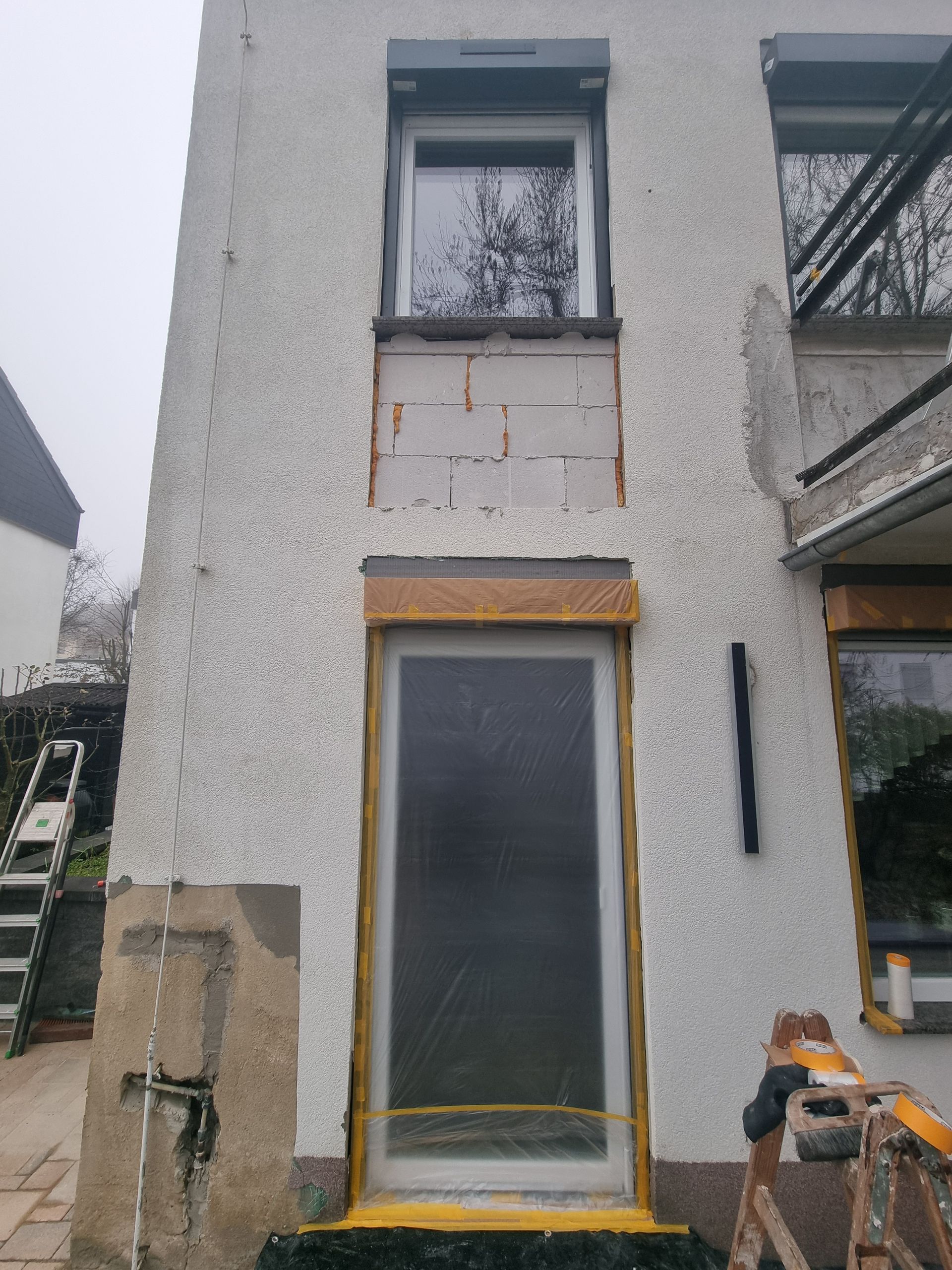 Hohes, weißes Haus mit gelb gerahmter Tür, kleinem Oberfenster und einem Gerüst auf der rechten Seite.
