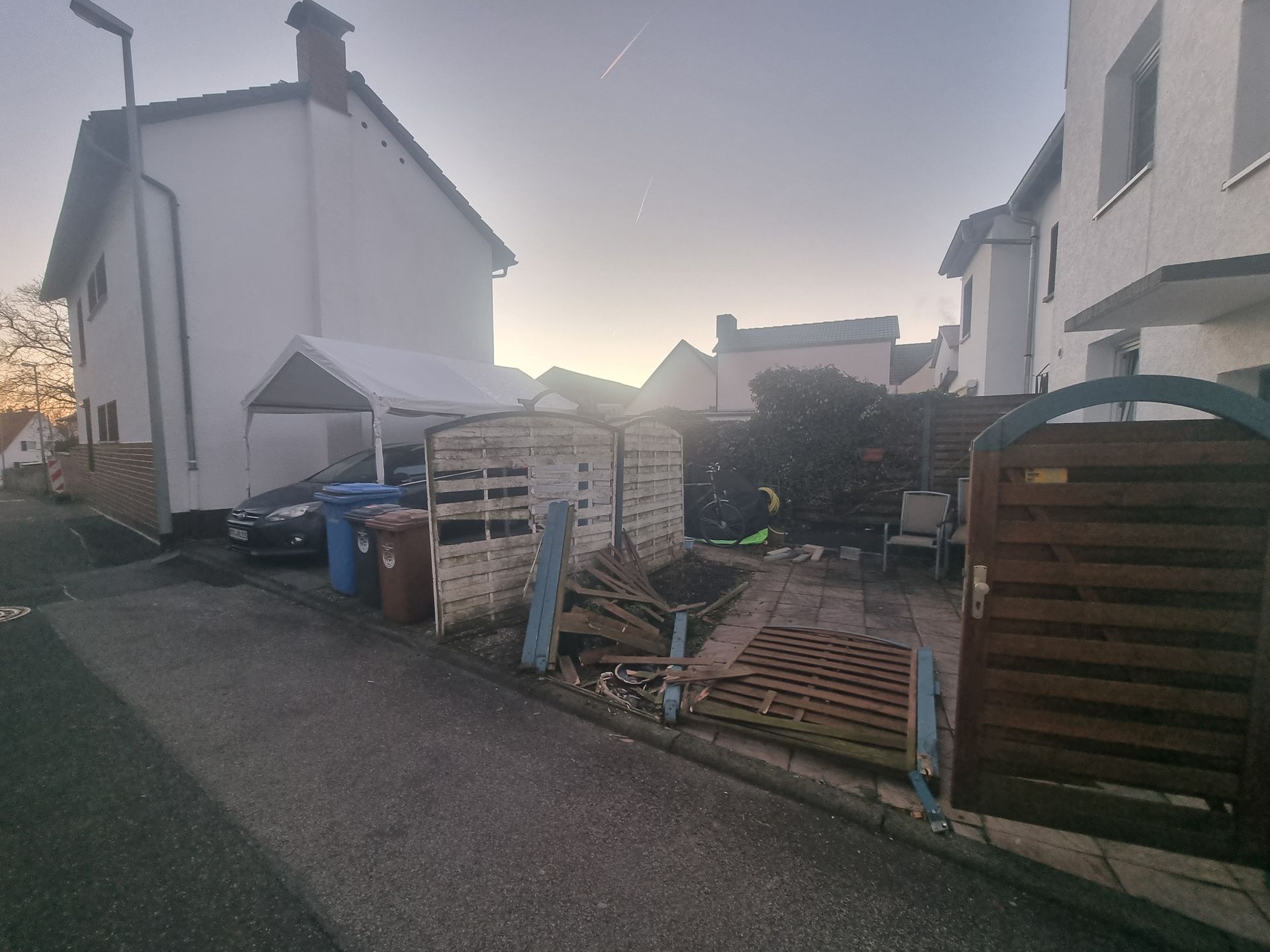 Eine schmale Wohngasse mit geparkten Autos, Mülltonnen, einem Zaun und einem kleinen Schuppen in der Abenddämmerung.