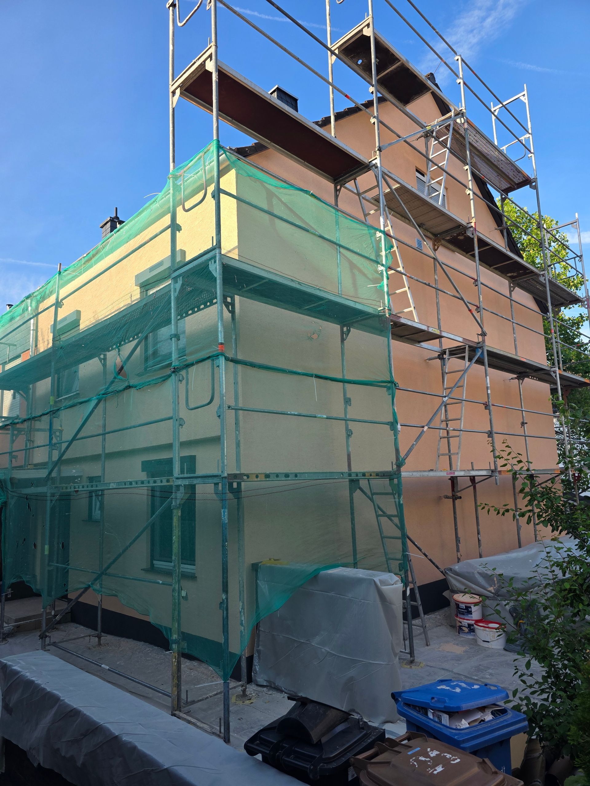 Ein im Bau befindliches Haus, eingehüllt in grüne Gerüstnetze, neben einem blauen Himmel