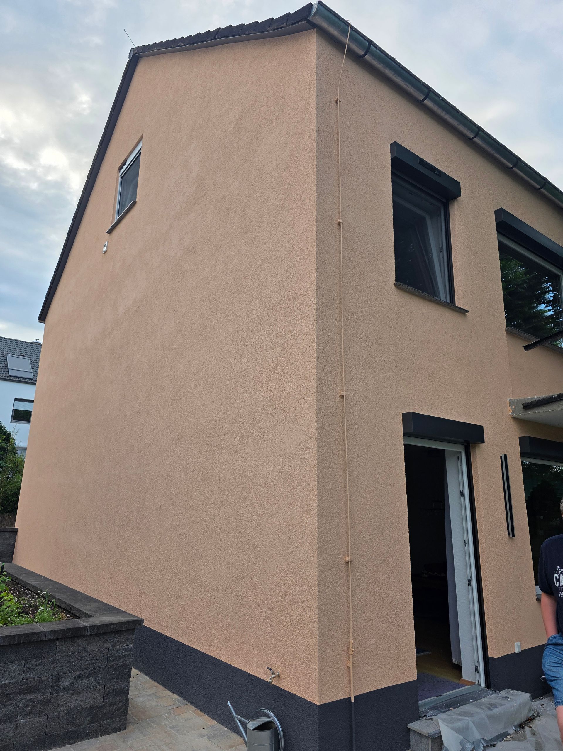 Eckansicht eines beigefarbenen Stuckhauses mit dunkel umrandeten Fenstern und einer Tür unter einem bewölkten Himmel