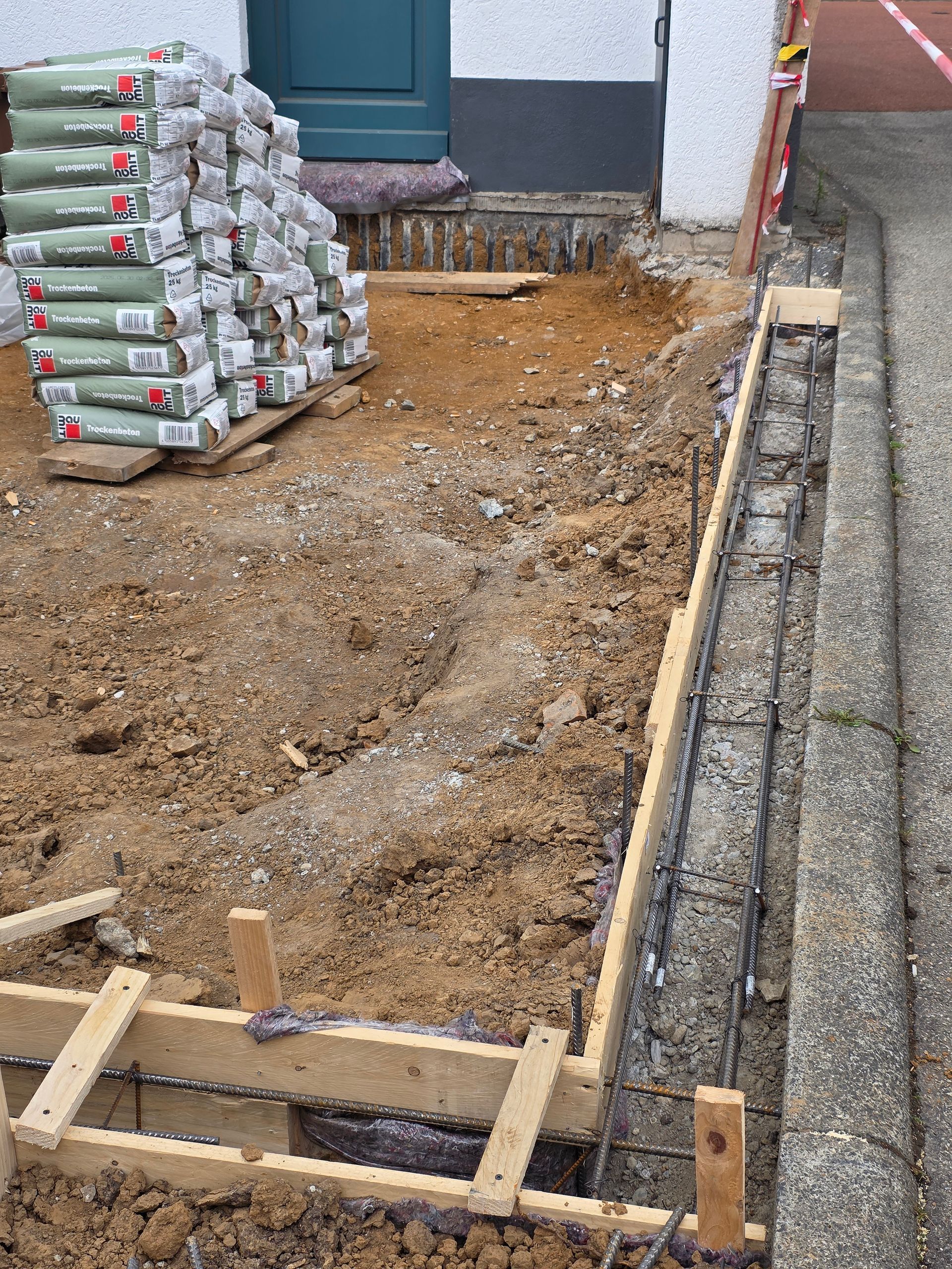 Baustelle mit Kiesbett, gestapelten Zementsäcken und Holzrahmen neben einer Hauswand
