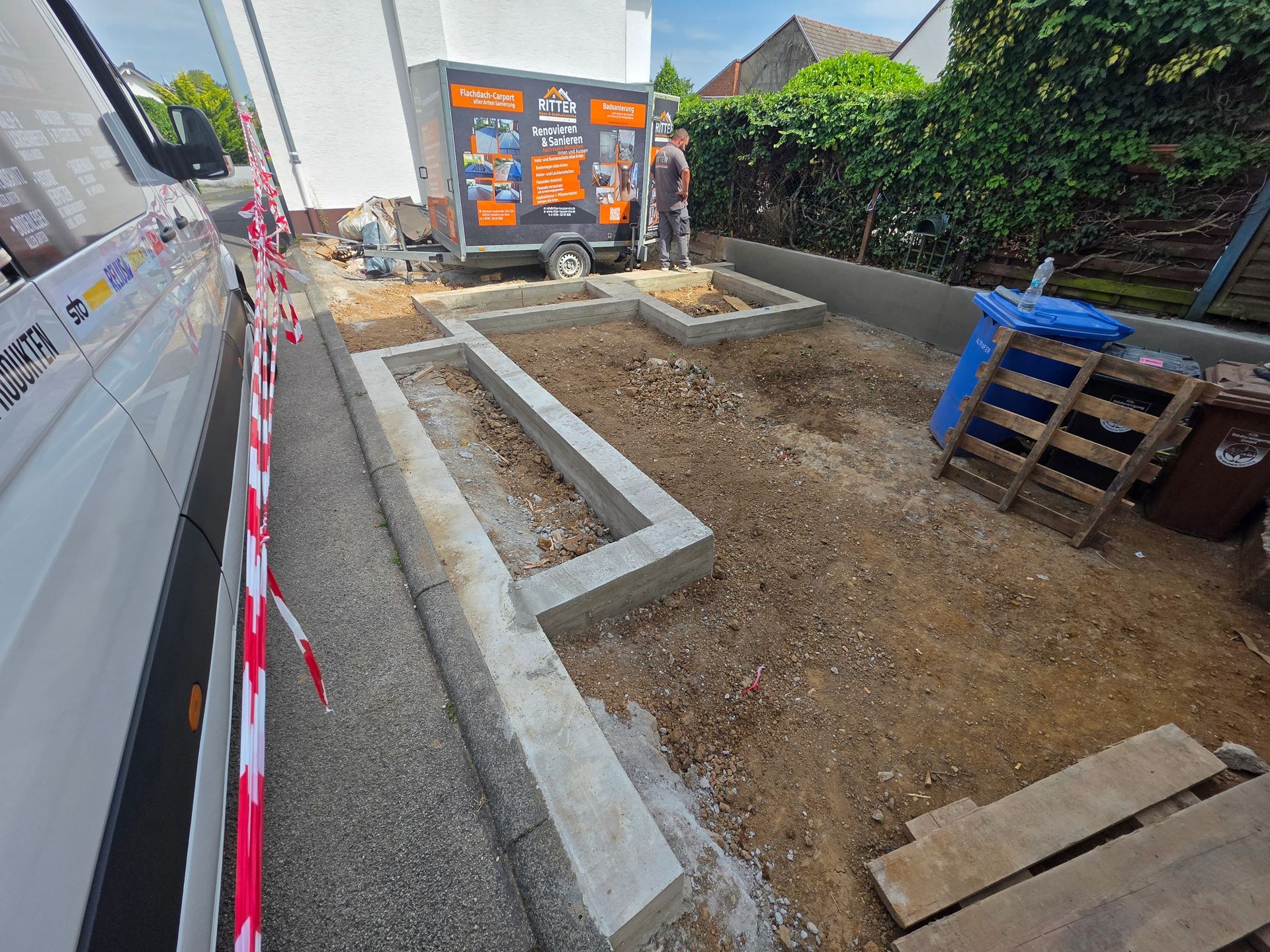 Freiluftbaustelle mit Betonfundamentwänden, Kiesboden und Baumaschinen in der Nähe eines Hauses.