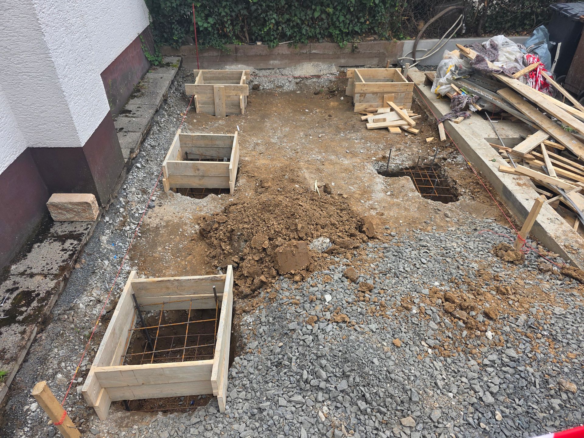 Baustelle mit Kies, Holzschalung und ausgehobenem Fundamentbereich neben einer Mauer