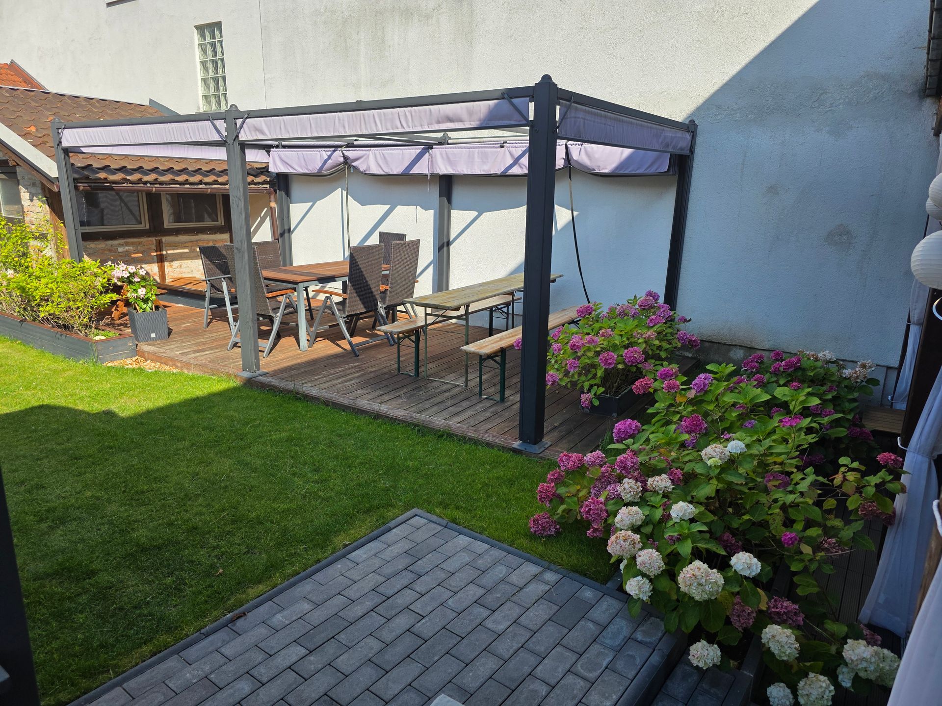 Hinterhofterrasse mit Pergola, Sitzgelegenheiten im Freien, Ziegelweg und blühenden Blumen neben einem Rasen.