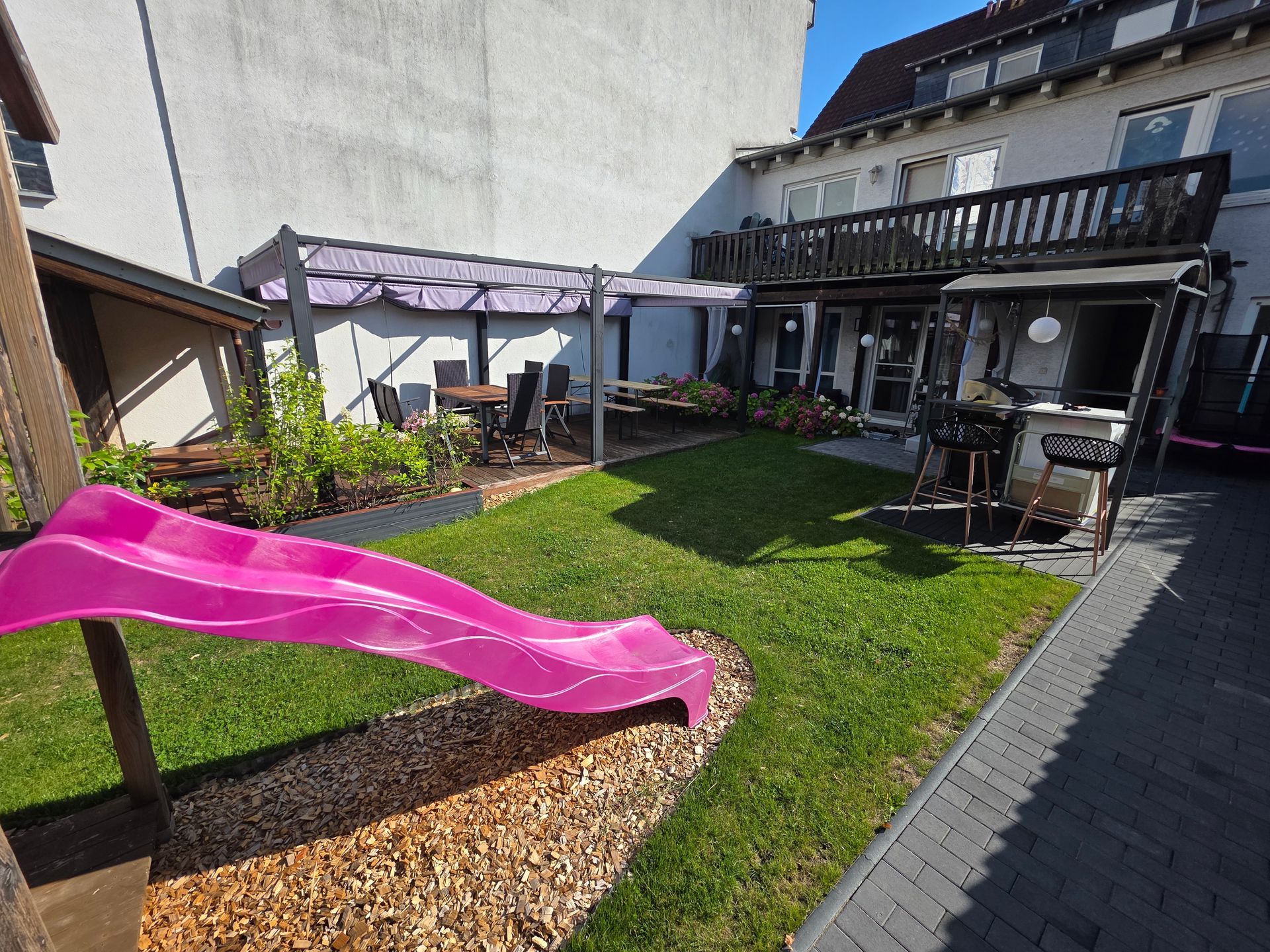 Hinterhofterrasse mit leuchtend pinkfarbener Rutsche, Pergola, Rasen und gepflastertem Gehweg neben einem Haus.
