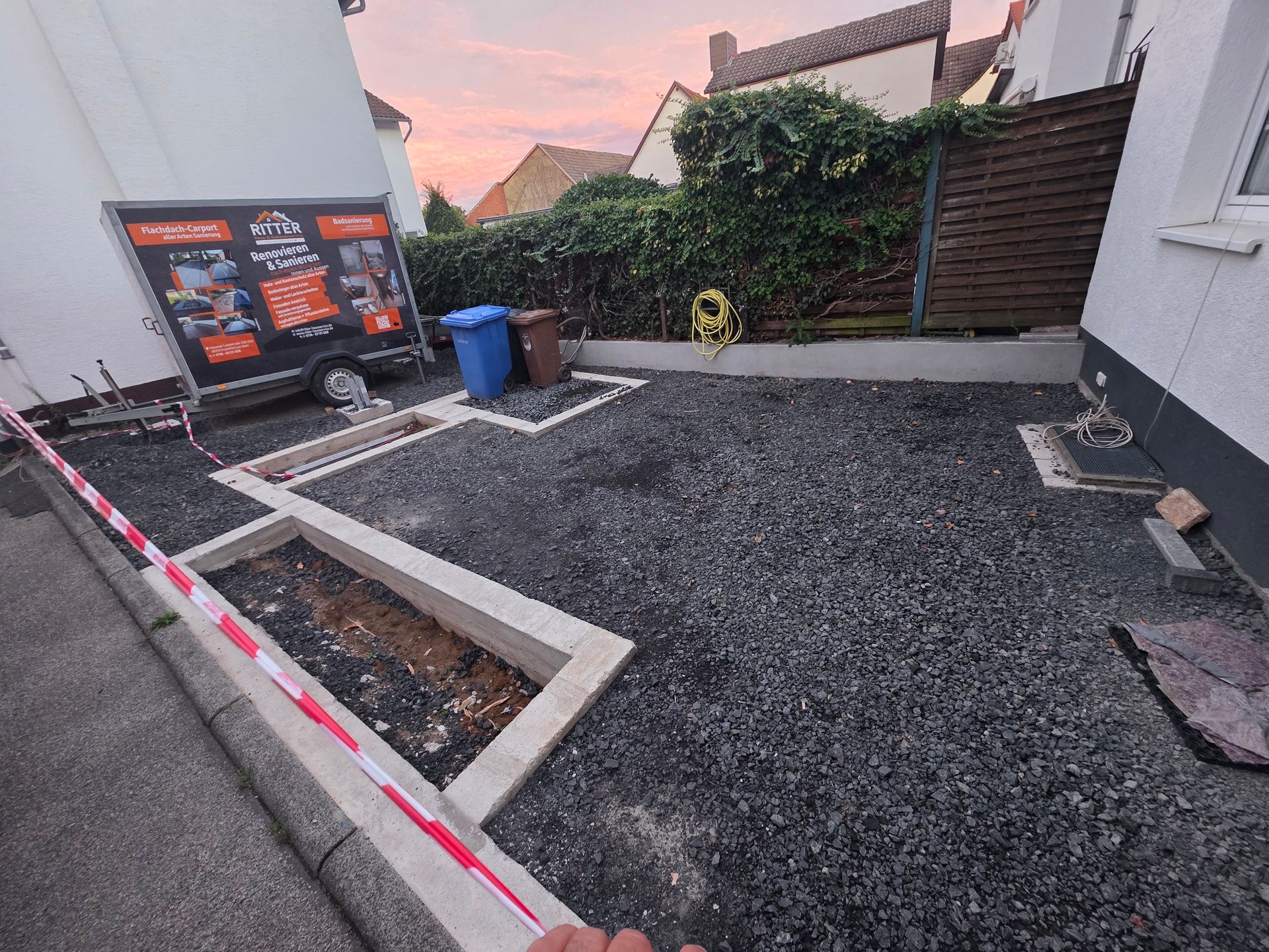 Eine mit Kies befestigte Auffahrt neben einem Haus mit erhöhten Betonrändern, einer blauen Mülltonne und einem Hinweisschild in der Nähe einer Hecke.